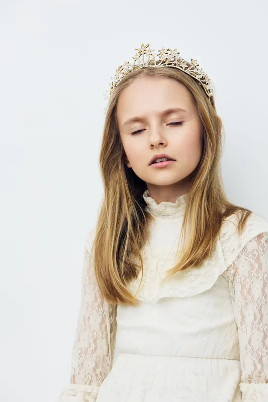 Young girl with a crown, wearing a white lace dress, eyes closed, conveying an ethereal and dreamy emotion against a soft white background