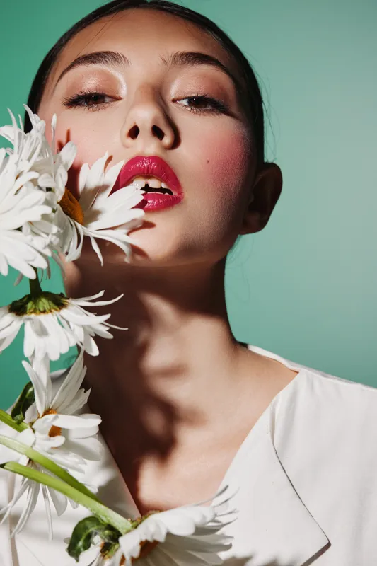 Close-up of a young female fashion model with bright pink lips holding white daisies, wearing soft shimmery eye makeup and pale blush on green background, beauty creative concept.