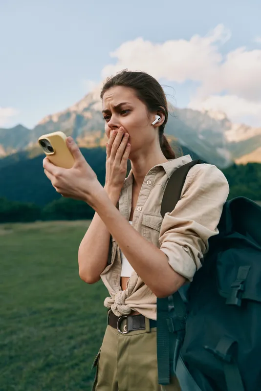 Surprised woman traveler outdoors checks her phone, wearing a backpack and beige shirt, mountains in the background, conveying curiosity and readiness.