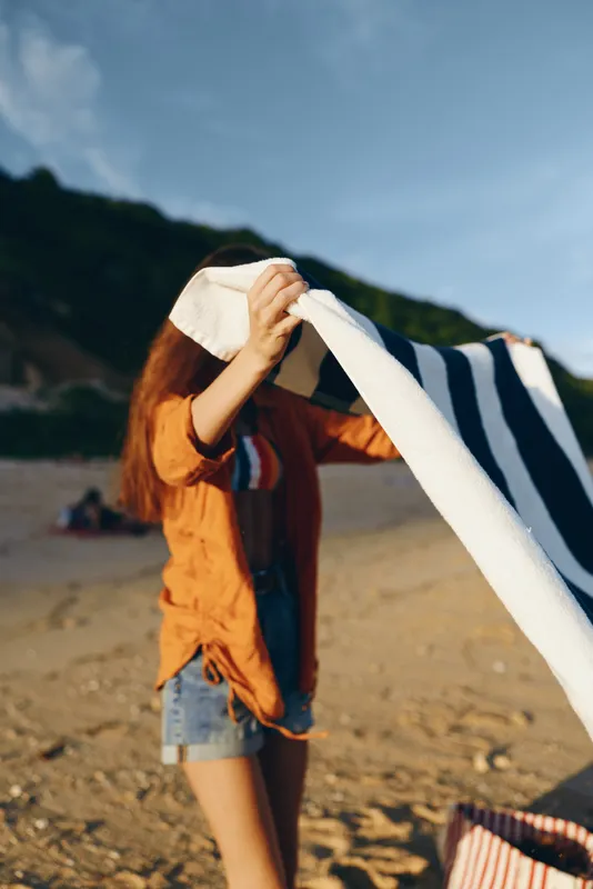 Woman on beach in summer holds striped towel near face, sunshine outdoors with travel mood, mountains behind, fashion casual lifestyle, bright smile, breeze relaxation, vacation portrait