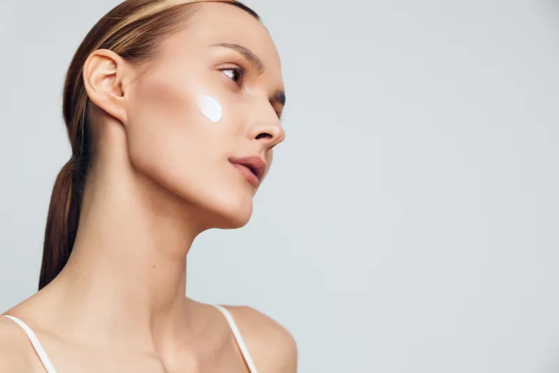 Young woman with clear skin applying white face cream on cheek. Side profile on light background. Concept of skincare, beauty routine, and healthy lifestyle. Soft natural lighting.