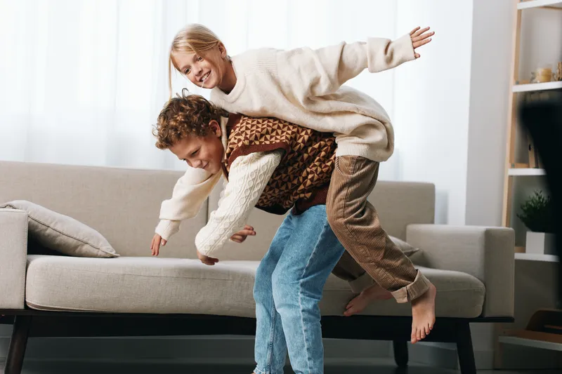 Playful moment of a man giving a child a piggyback ride on a cozy living room sofa, bright natural light, warm tones, candid happiness, and family bonding.