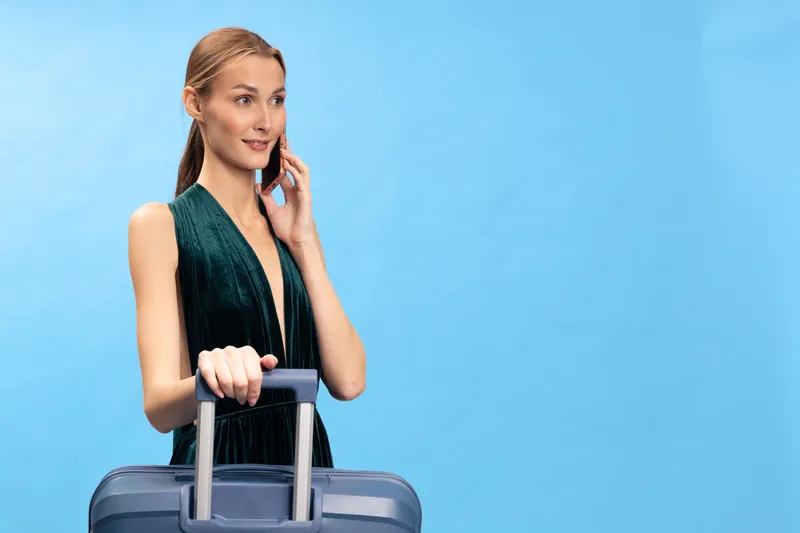 Young woman in green dress talking on phone and holding suitcase handle on blue background. Concept for travel, vacation, communication, and lifestyle with luggage and casual fashion.