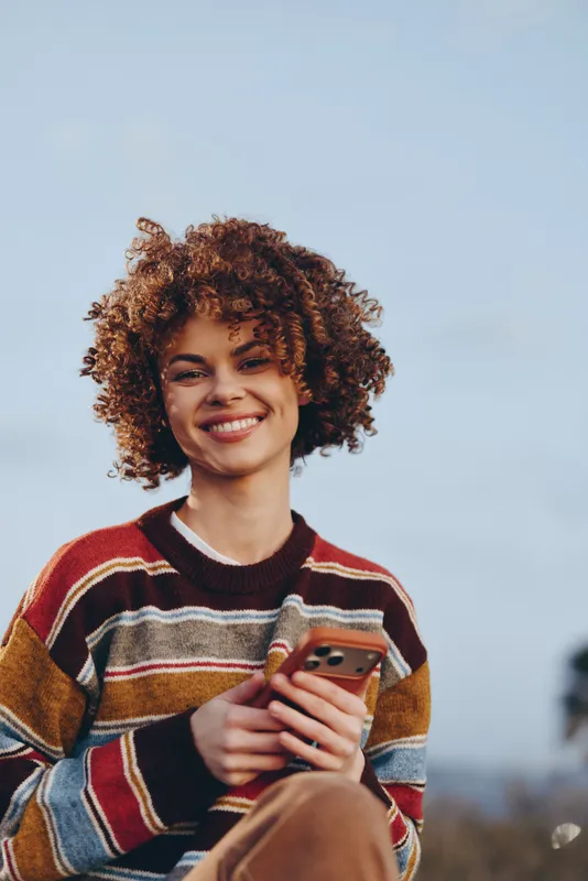 Young woman lifestyle outdoors, with a smile, curly hair, striped sweater, holding smartphone in her hands, sunny sky background, relaxed casual moment, soft light, natural colors