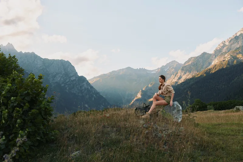 Woman lifestyle traveler sitting on rock in mountain landscape, enjoying nature, casual outfit, peaceful moment, sunny weather, outdoor adventure