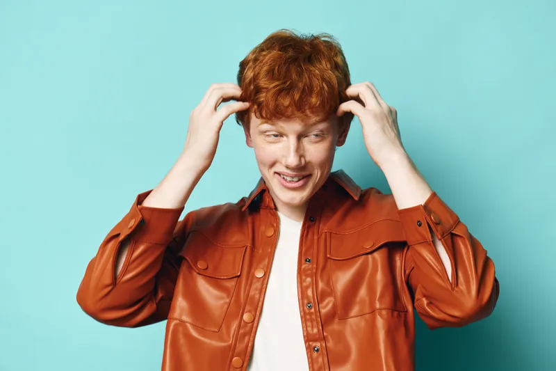 Young man with red hair wearing brown leather jacket touches head smiling and looking down on light blue background. Casual style, portrait, emotions and hairstyle concept in studio.