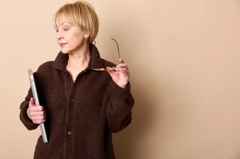 Mature blonde woman holds a book and reads, tired frustrated expression, indoor studying mood, education concept, beige background, copy space, slightly looking away from camera.
