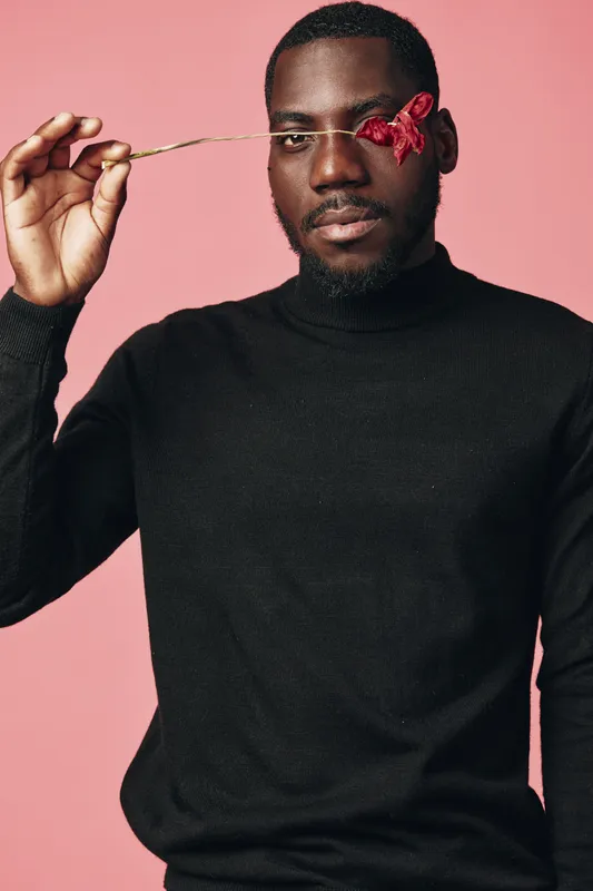African man with dark skin and beard holding red flower near eye in creative conceptual style. Portrait with pink background and black sweater for visual artistic expression and fashion.