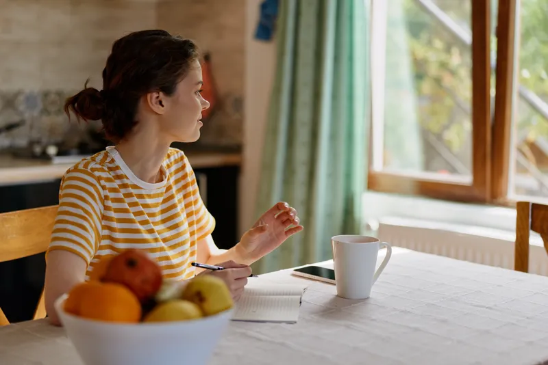 Woman in a striped shirt sits at a bright kitchen table, gazing through a window. Morning light highlights a bowl of fruit and a mug, conveying everyday lifestyle and home comfort.