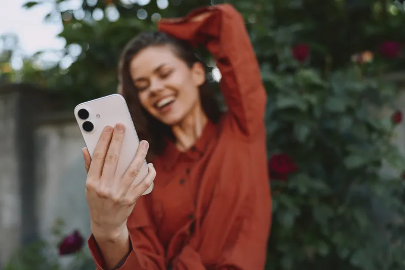 Young woman with brunette hair takes a cheerful selfie in a blooming garden, wearing a red blouse, capturing a candid and joyful moment
