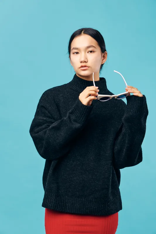 Young woman with dark hair and fair skin wearing black sweater holding white sunglasses against blue background. Casual portrait of fashion style and confident female model in studio setting.