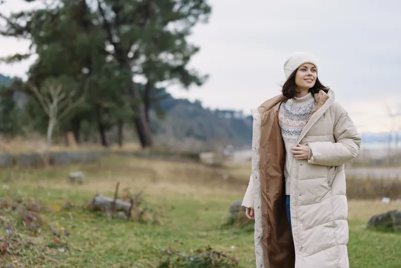 Woman in a long beige winter coat stands in an open field with trees and hills, smiling softly, wearing a knit sweater and beanie for a calm seasonal mood outside