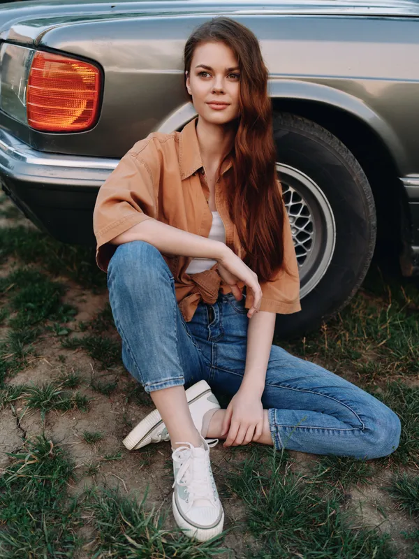 Young woman sitting beside vintage car in casual summer outfit, showcasing relaxed mood with natural beauty and denim fashion.