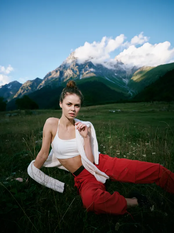 Woman sits on a grassy meadow surrounded by mountains, wearing a white crop top and red pants, with a light cardigan, embracing nature and outdoor fashion
