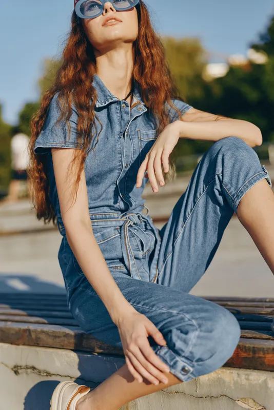 lifestyle woman in modern boho-western denim street style sitting outdoors, film color tones, casual summer fashion, relaxed pose with sunglasses