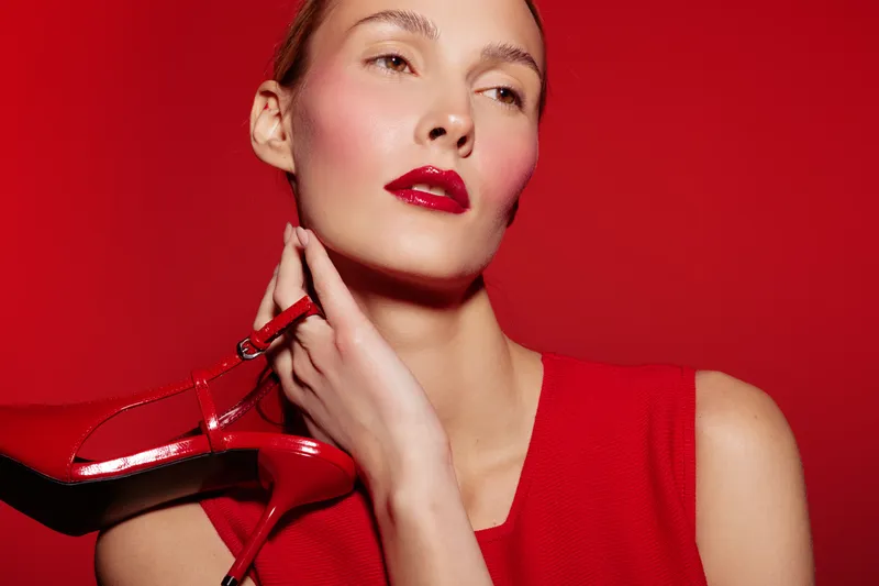 Young woman in red dress holds red high heel shoe near face on red background. Fashion concept with bold makeup and stylish accessories in studio lighting.