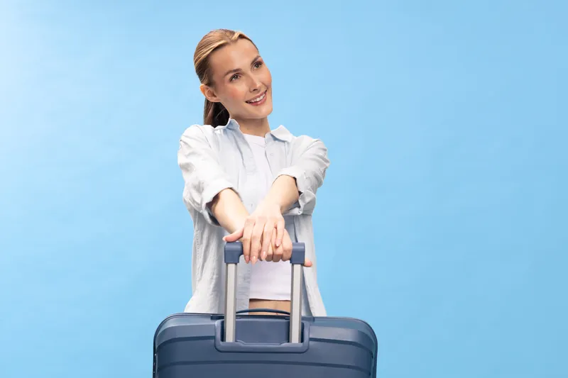 Young woman with suitcase smiles happily against blue background. Travel concept with positive emotions and casual clothing. Ready for vacation or business trip with luggage.