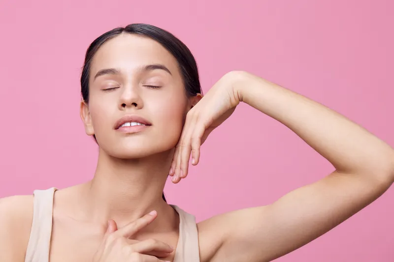 Beautiful young woman with closed eyes smiling gently against a soft pink background, showcasing natural beauty and tranquility in a fashion forward style