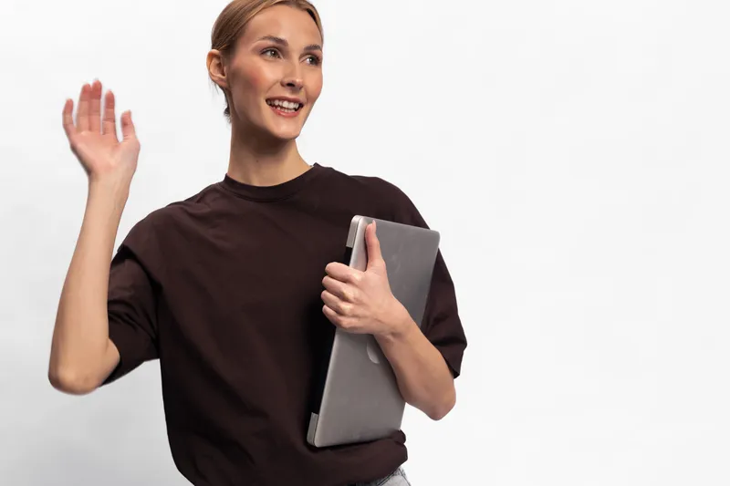 Smiling young woman holding laptop waves hand greeting someone. Casual female in dark shirt isolated on white background. Concept of communication, technology, and friendly gesture.