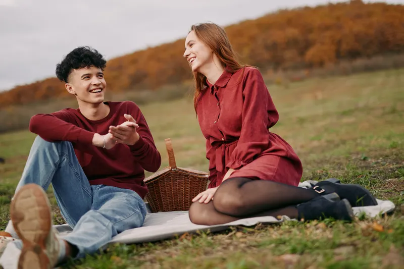 A candid outdoor portrait of a young couple sharing laughter during a picnic on a meadow, capturing authentic emotion and natural interaction that highlights genuine connection and relaxed chemistry.