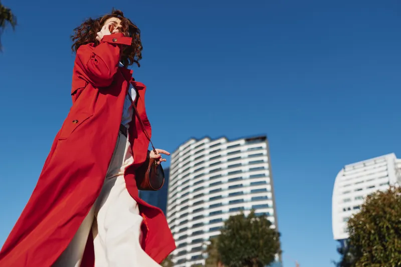 A young woman in a bright red trench coat walks outdoors among modern buildings, looking up with confidence as the blue sky shines above, creating a bold city mood.