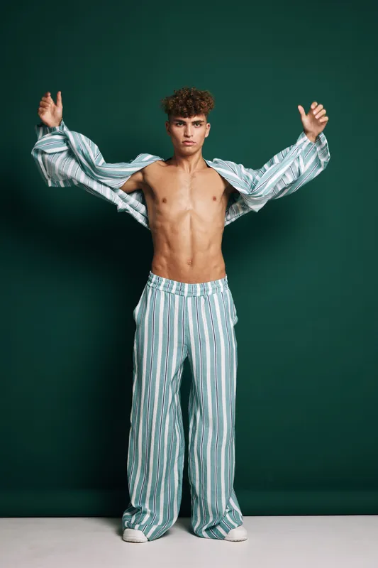 Young man with curly hair and toned body wearing striped wide pants and matching open shirt posing with hands raised on dark green background for fashion and style photography.