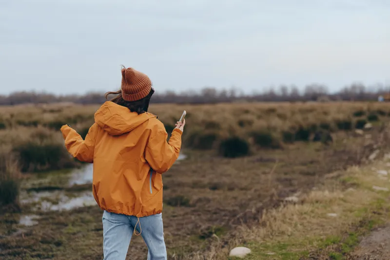 Woman outdoor lifestyle in open field, wearing yellow jacket and knit hat, standing with peace gesture, cloudy daylight, relaxed posture, rural meadow landscape, natural colors