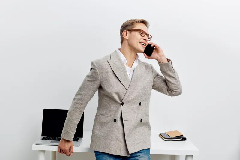 Confident young man in smart casual attire talking on smartphone in bright office environment isolated on plain background. Business communications, professional, cheerful, modern style, male professi
