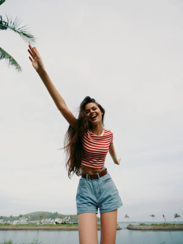 Joyful young woman outdoors wearing a striped crop top and denim shorts, arms raised and smiling, capturing freedom and playful energy against a bright sky near the water.