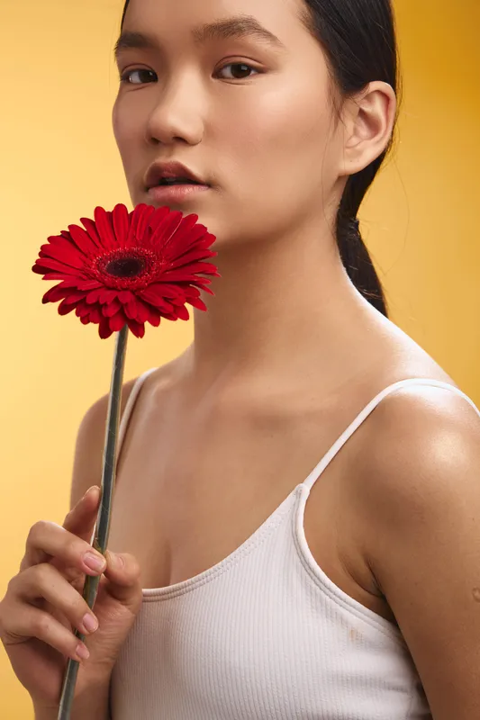 asian woman photographed in a soft studio setting, holding a red flower near her lips, bright yellow background highlights her serene expression and natural beauty.
