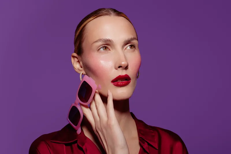 Stylish woman with slicked hair and glossy red lips holds pink sunglasses near face on purple background. Fashion portrait with vibrant colors and modern accessories concept.