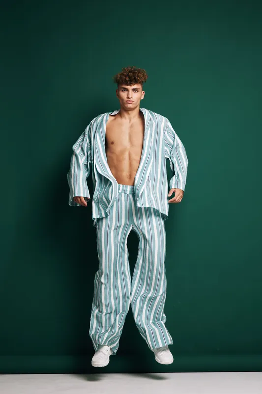 Young man with curly hair wearing striped loose pajamas standing against dark green background with open shirt and white sneakers in studio.