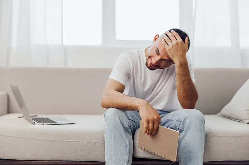 Young man sitting on a sofa with a notebook, looking pensive and stressed, surrounded by soft natural light. Ideal for showcasing modern life challenges and mental health themes.
