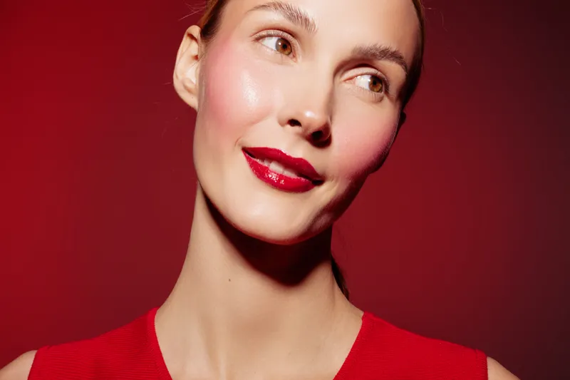 Close-up portrait of a smiling woman with glossy red lips and smooth skin wearing a red top against a red background. Concept of beauty, makeup, and fashion with vibrant colors.