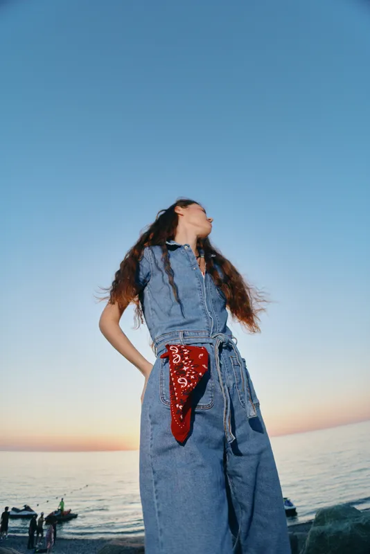 lifestyle woman in modern boho-western denim outfit with long hair and red bandana near seaside under clear blue sky, film color effect