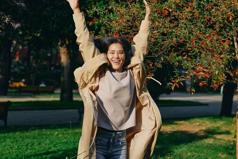 Joy, happiness, girl outdoors, coat, autumn, park, movement, celebration, energy, sunlit scene with a smiling youth enjoying outdoor play in a casual jacket amid trees and grass
