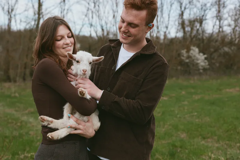 A young couple holds a baby goat together in a green field while smiling and enjoying a casual moment outdoors in cozy autumn clothes.