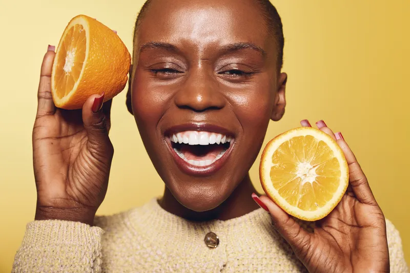 Happy dark skinned African woman holds two halves of orange fruit near face, smiling with closed eyes on yellow background. Concept of healthy lifestyle, vitamin C, and natural beauty.