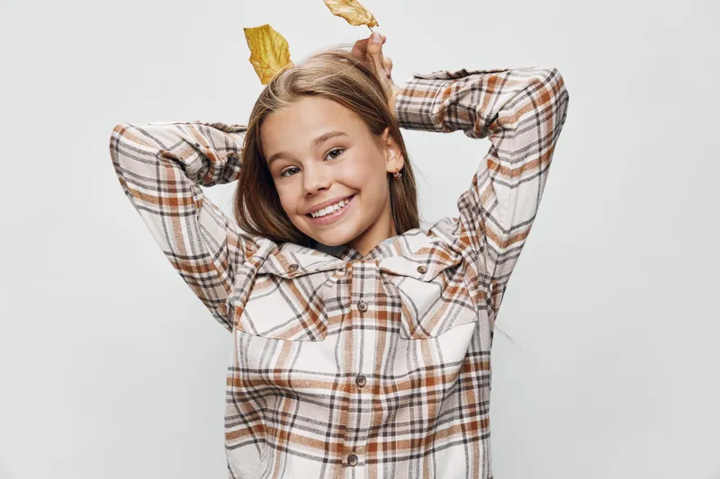 Smiling girl posing with autumn leaves in hands wearing checked shirt on white background for seasonal smiling portrait and childhood happiness outdoors.