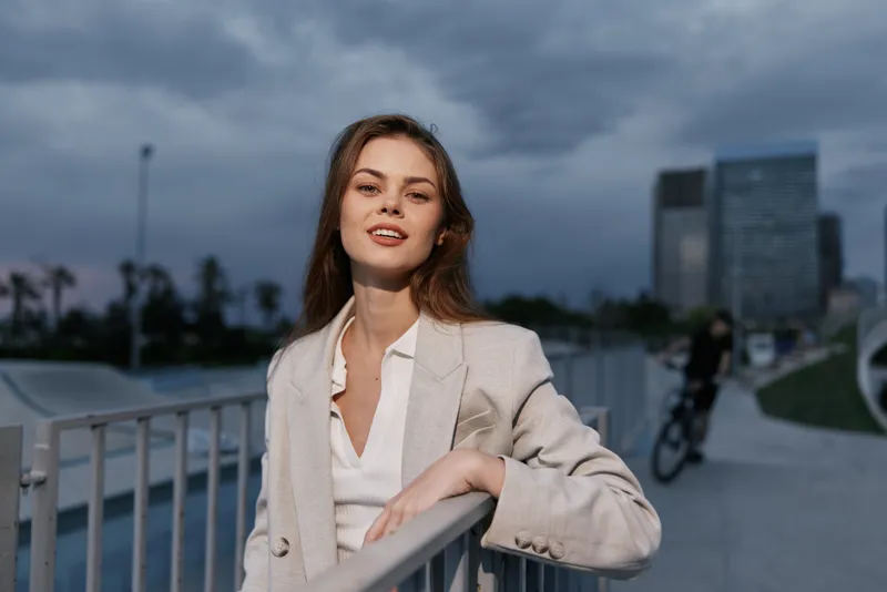 Young woman in beige blazer confidently posing outdoors with urban cityscape background cloudy sky business fashion style