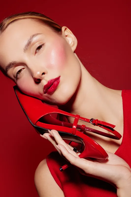 Close-up of a young woman with glossy red lips holding a red high heel shoe near her face against a red background. Concept of fashion, beauty, and style with vibrant colors.