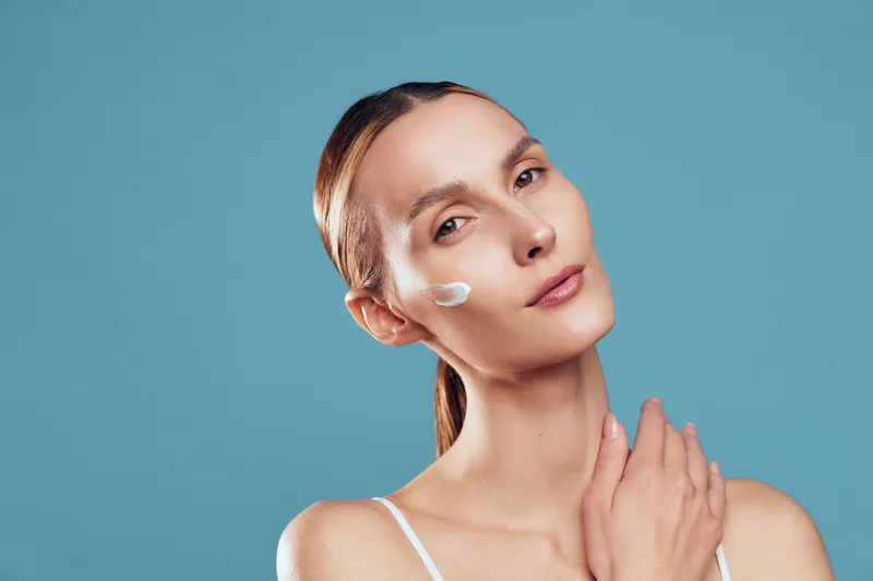 Woman skincare portrait applying face cream and gentle lotion on cheek against blue background, beauty close up of calm model with hands near face, fresh clean look today