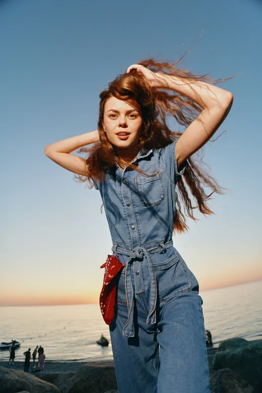 Lifestyle woman in modern boho-western denim street style with flowing hair and sunset sea background, captured in warm film color tones for a natural look.