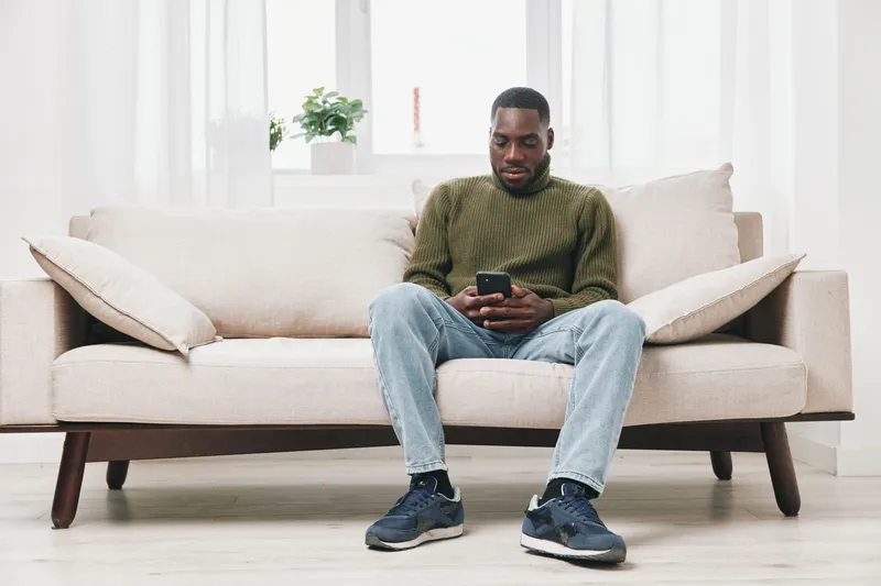 Young African American man sitting on a sofa, using a smartphone, dressed casually in a green sweater and jeans, conveying relaxation and modern lifestyle
