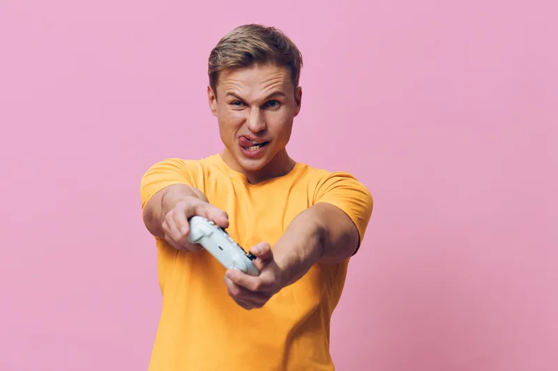 Angry young man with short hair wearing yellow t-shirt holding game controller, expressing frustration or aggression, on pink background, studio portrait, emotional, colorful, casual style. People lif