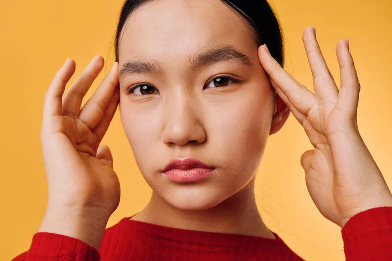 Woman with clear skin and natural makeup posing against orange background touching temples with fingers, wearing red sweater, looking at camera with calm expression indoors.