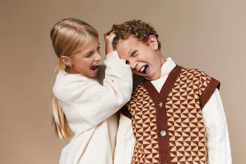 Two young children laugh and play together, sharing a playful moment in cozy knit sweaters against a warm neutral backdrop, capturing joy, friendship, and lighthearted energy.