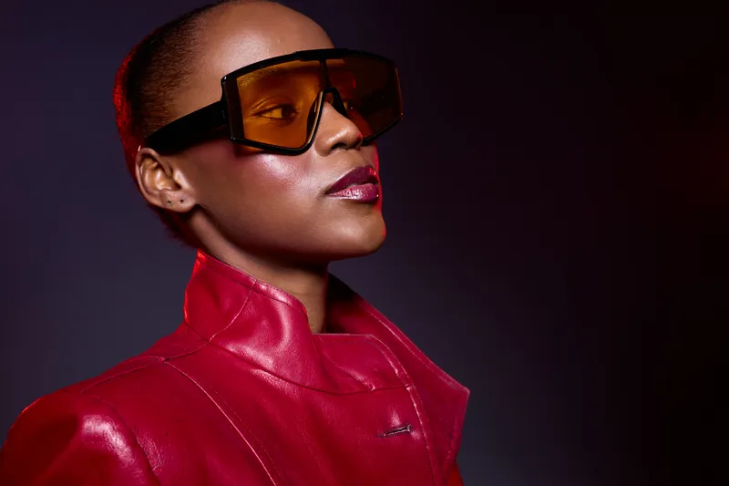 Confident black woman with dark skin wearing large brown sunglasses and a red leather jacket, posing against a dark background. Concept of fashion, style, and modern elegance.