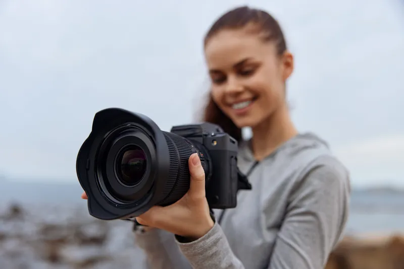 Young woman with a camera at the beach, wearing a gray hoodie, smiling joyfully towards the ocean, capturing memories in a beautiful coastal setting