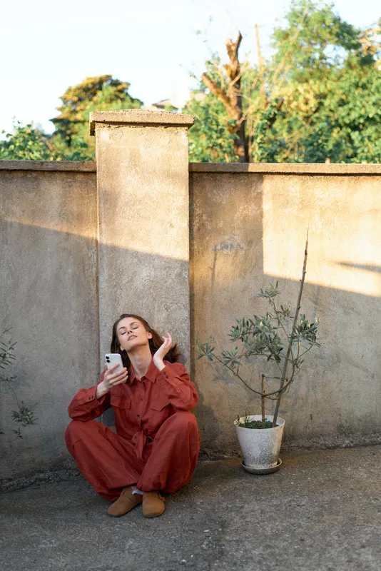 Casual woman in red jumpsuit sits against a wall outdoors, looking at her phone. Street style scene with warm sunlight, simple surroundings, relaxed mood, everyday life and self expression